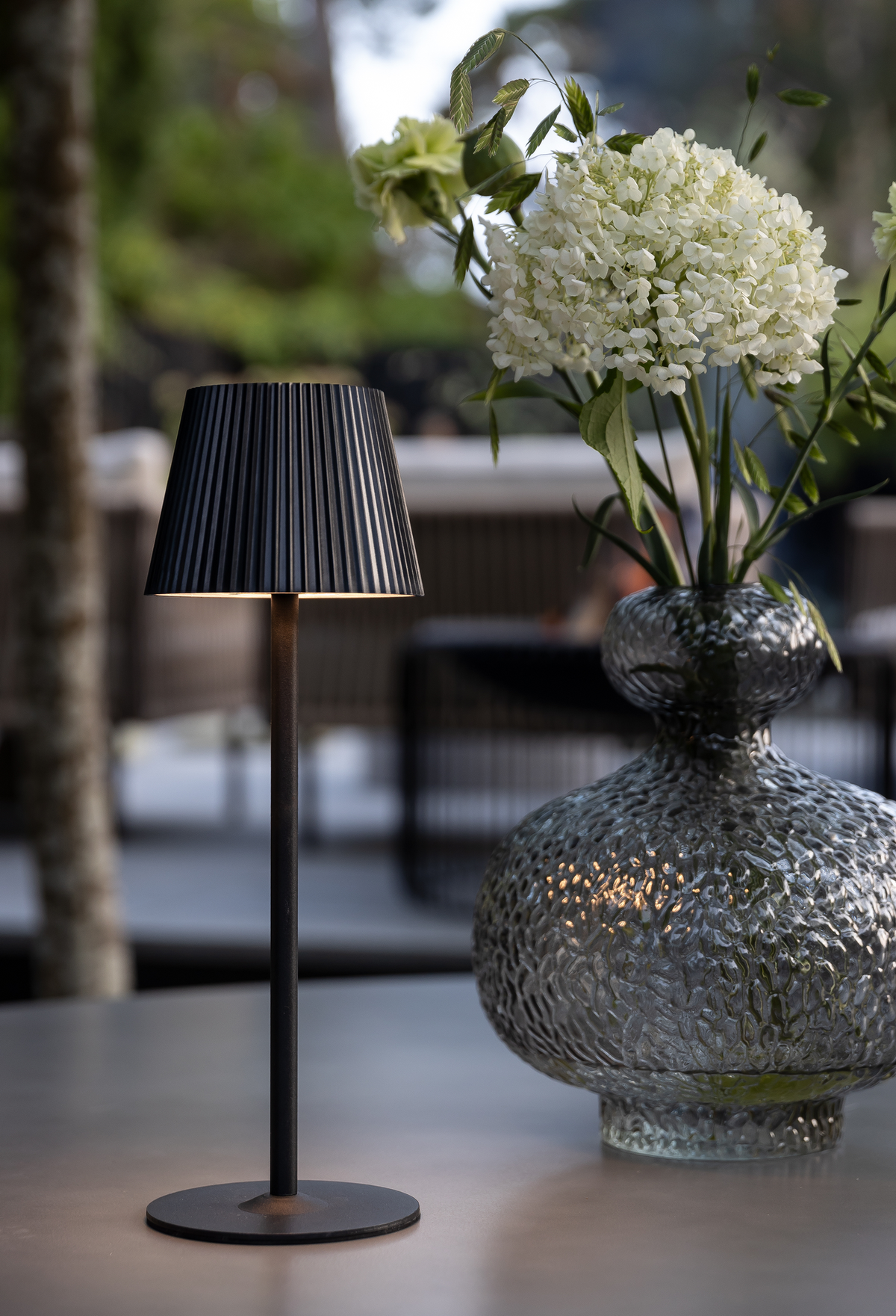 Sort bordlampe med plisseret skærm ved siden af en rillet glasvase med hvide hortensia på en terrasse.