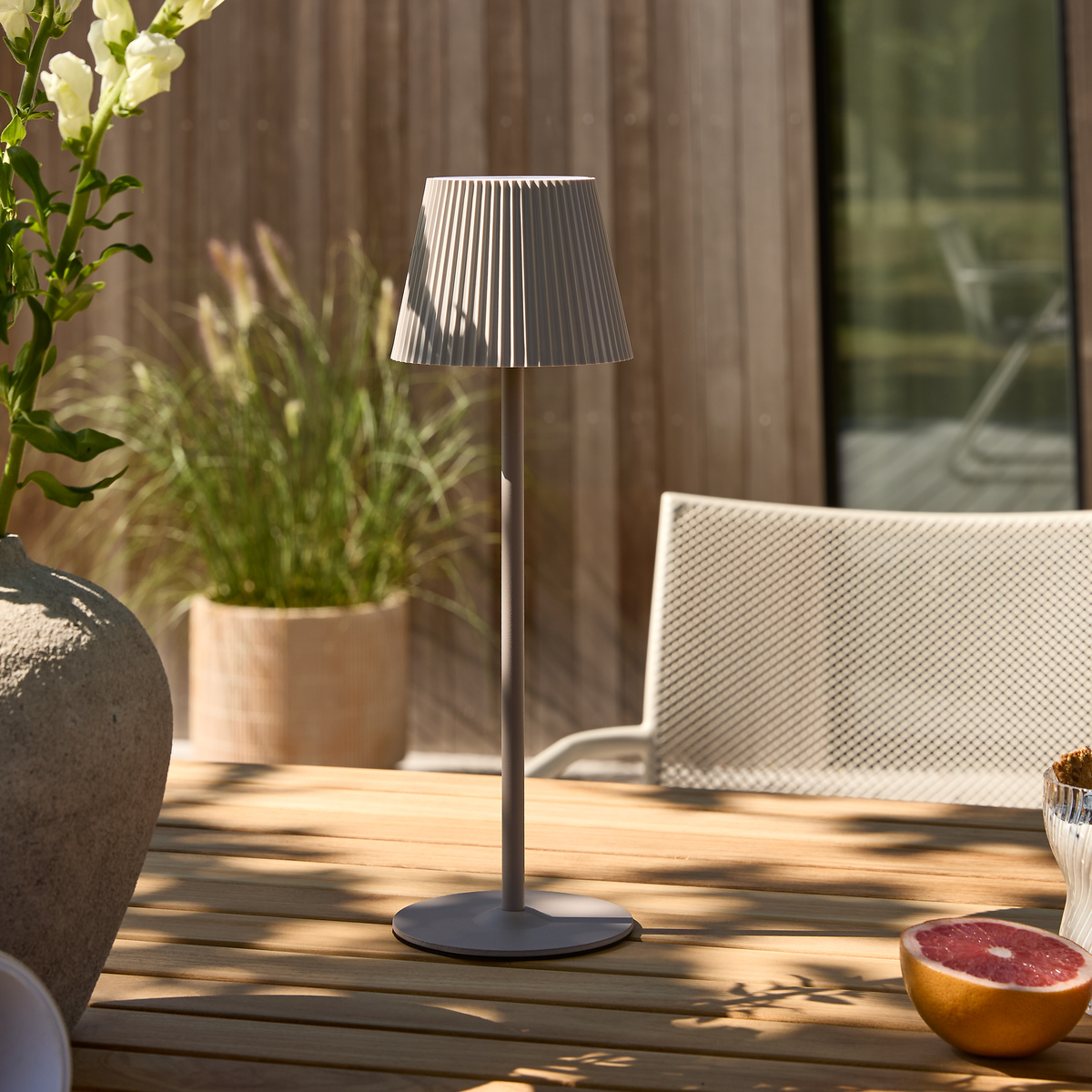 Bærbar udendørs bordlampe i beige med plisseret skærm på et træbord på terrassen, omgivet af krukkeplanter og en netstol i solskin.