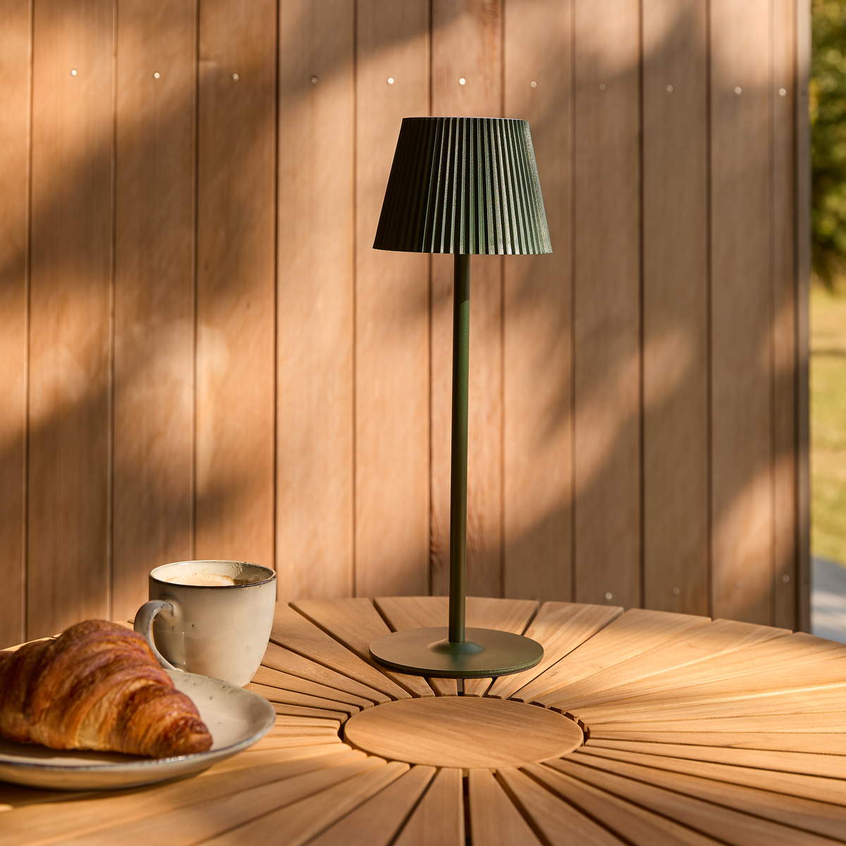 Grøn bærbar bordlampe på rundt træbord udendørs ved siden af en kop kaffe og en croissant i varm morgensol.