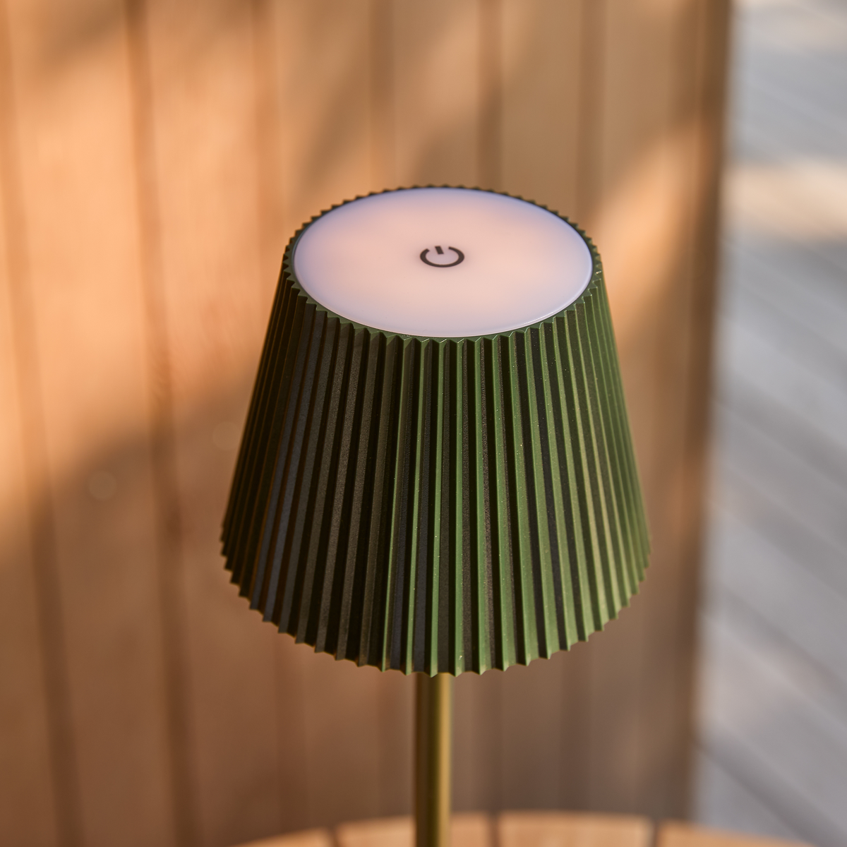 Nærbillede af en grøn bærbar bordlampe med plisseret skærm og en tænd/sluk‑symbol på toppen i varmt dagslys.