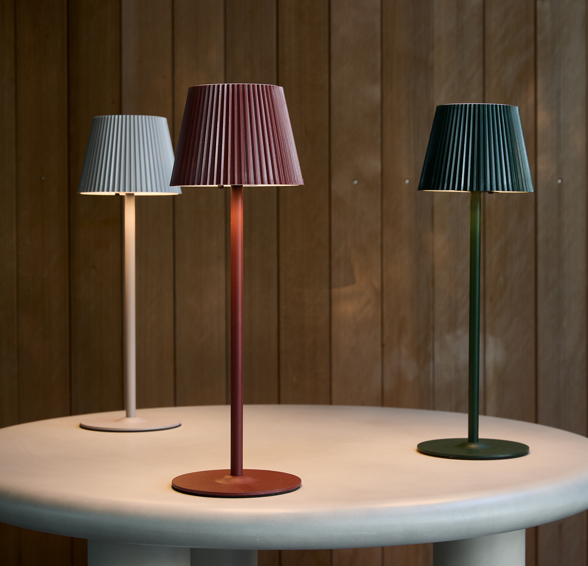 Tre slanke bordlamper med plisserede skærme i grå, rød og grøn står på et rundt lysegråt bord foran en træpanelvæg.