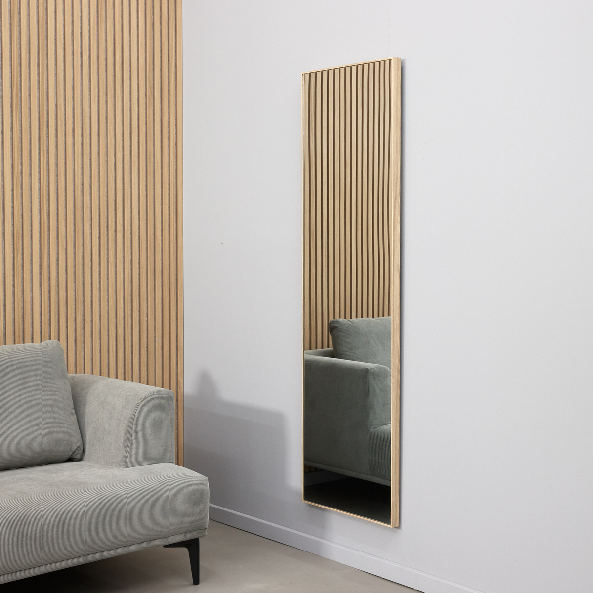 Smalt rektangulært vægspejl med lys træramme ved siden af en grå sofa og væg med trælameller i et minimalistisk stuemiljø.