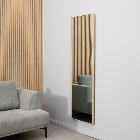 Smalt rektangulært vægspejl med lys træramme ved siden af en grå sofa og væg med trælameller i et minimalistisk stuemiljø.