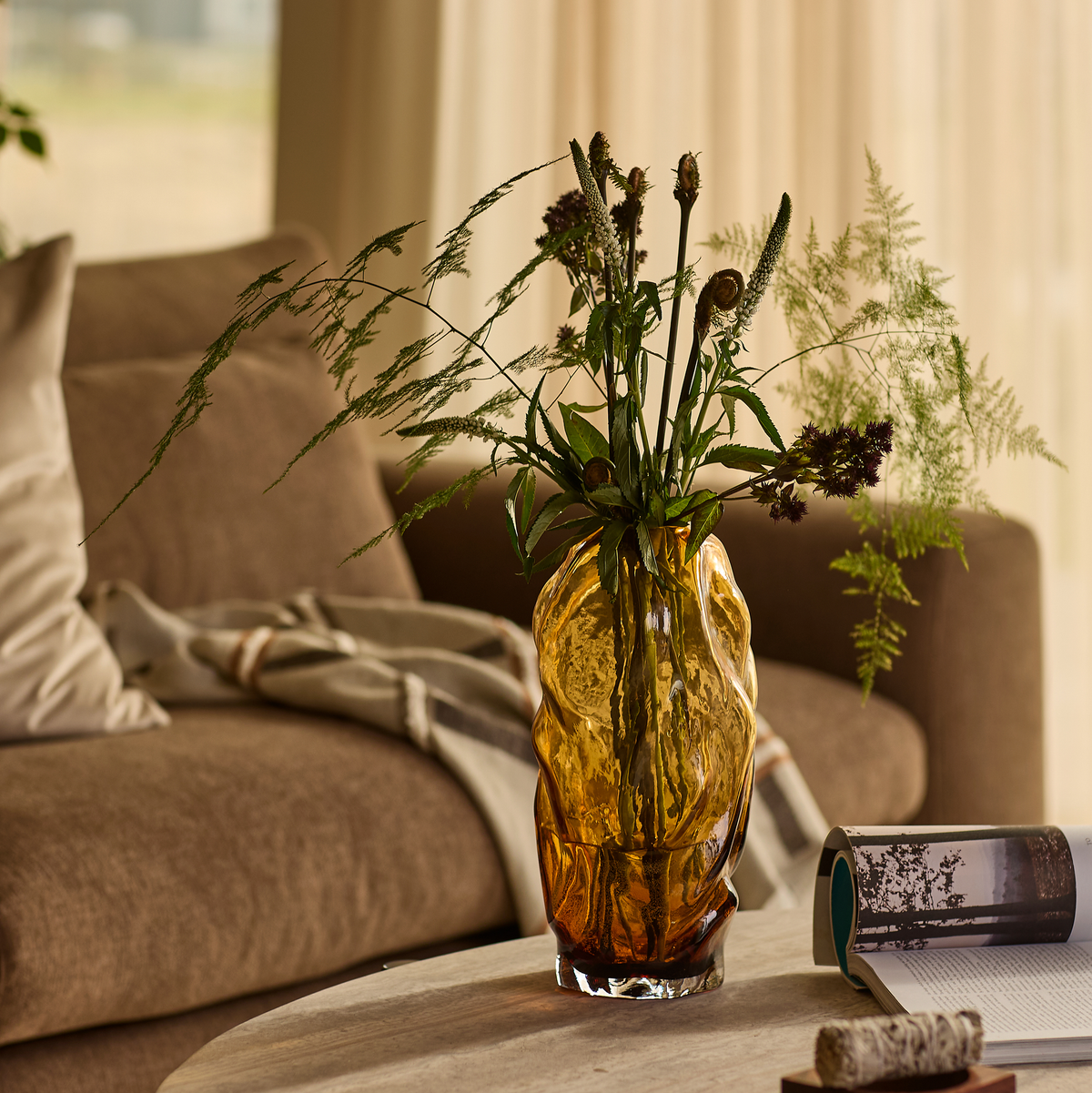 Ravfarvet skulpturel glasvase med grønne grene på et sofabord i en varm stue med sofa i baggrunden.
