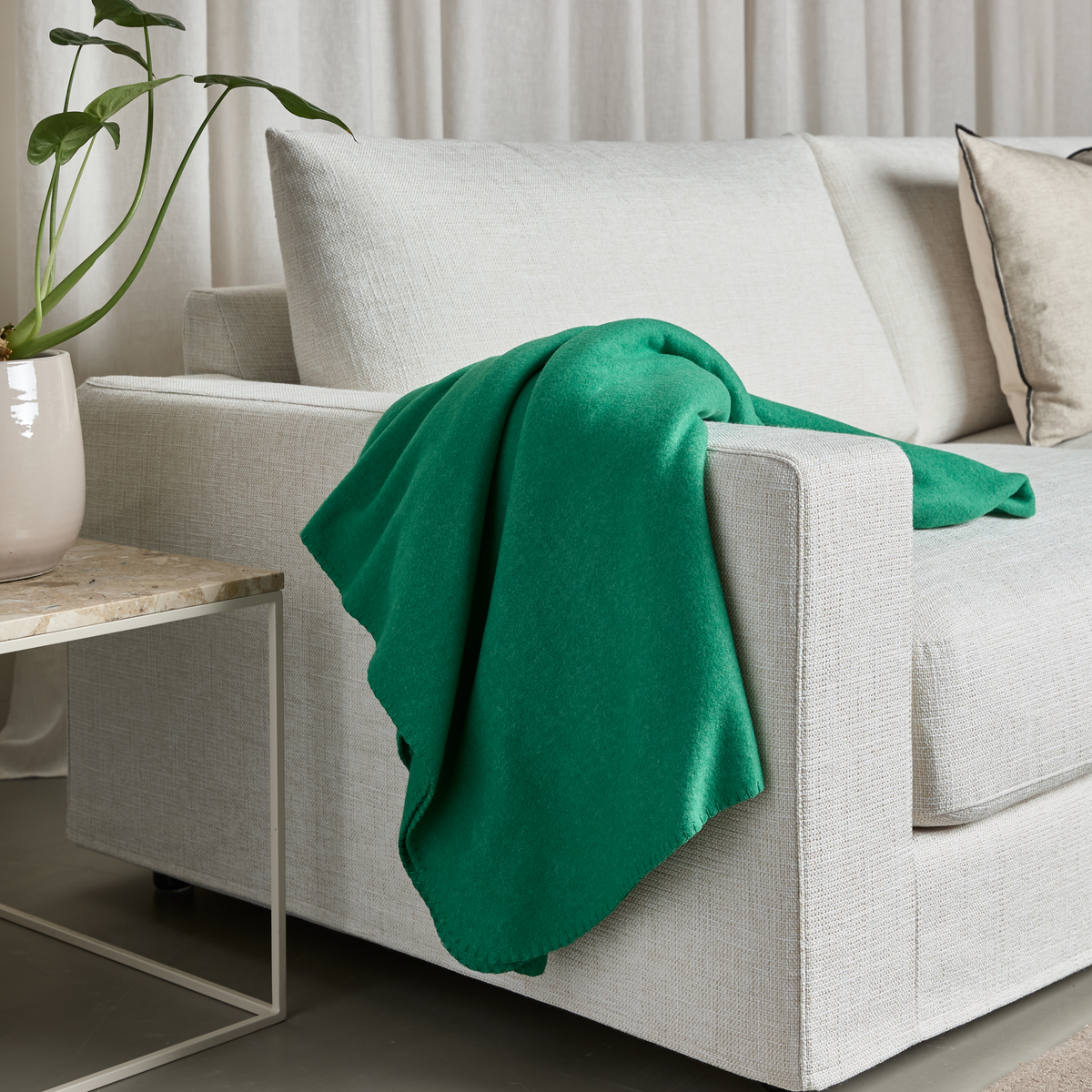 Lys beige sofa med grøn plaid over armlænet ved siden af et sidebord med vase og grøn plante.