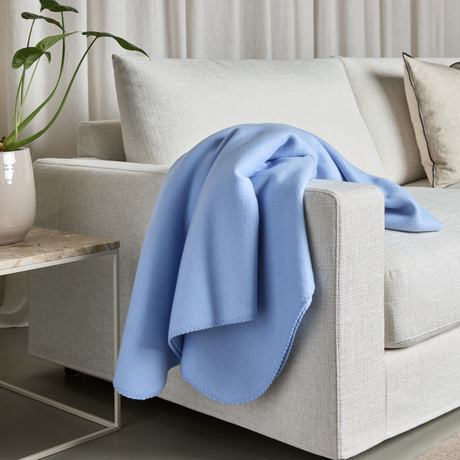 Lyseblåt plaid lagt over beige sofa med neutrale puder, ved siden af en potteplante på et sidebord.
