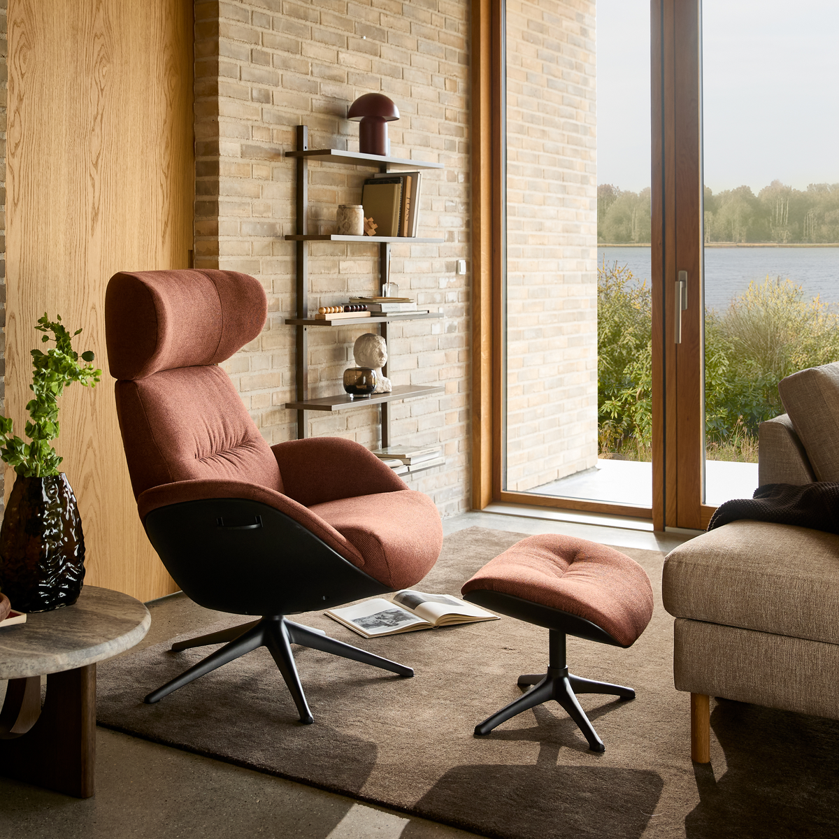 Rustrød lænestol med matchende skammel i en moderne stue, store vinduer med udsigt til sø, væghylde med lampe og bøger, beige sofa og brun tæppe.