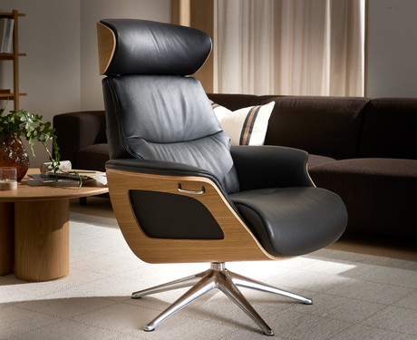 Sort læderlænestol med træskal og krom drejefod i en stue med sofa og sofabord i blødt dagslys.
