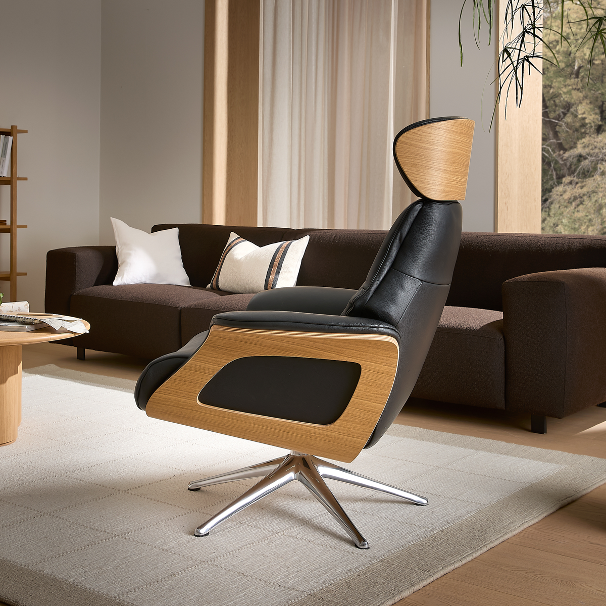 Moderne loungestol i sort læder med trædetaljer og metalfod i en lys stue med brun sofa, puder, gardiner, tæppe og planter.