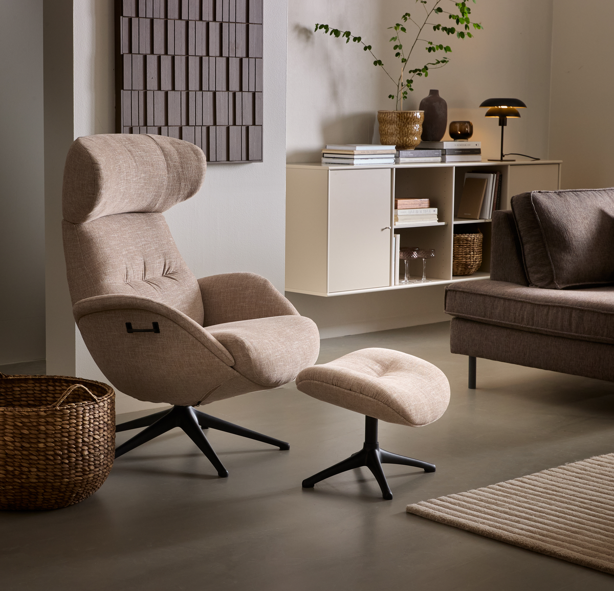 Beige loungestol med matchende fodskammel i en stue med vægmonteret hylde, bøger, bordlampe, plante, sofa og fletkurv i neutrale toner.