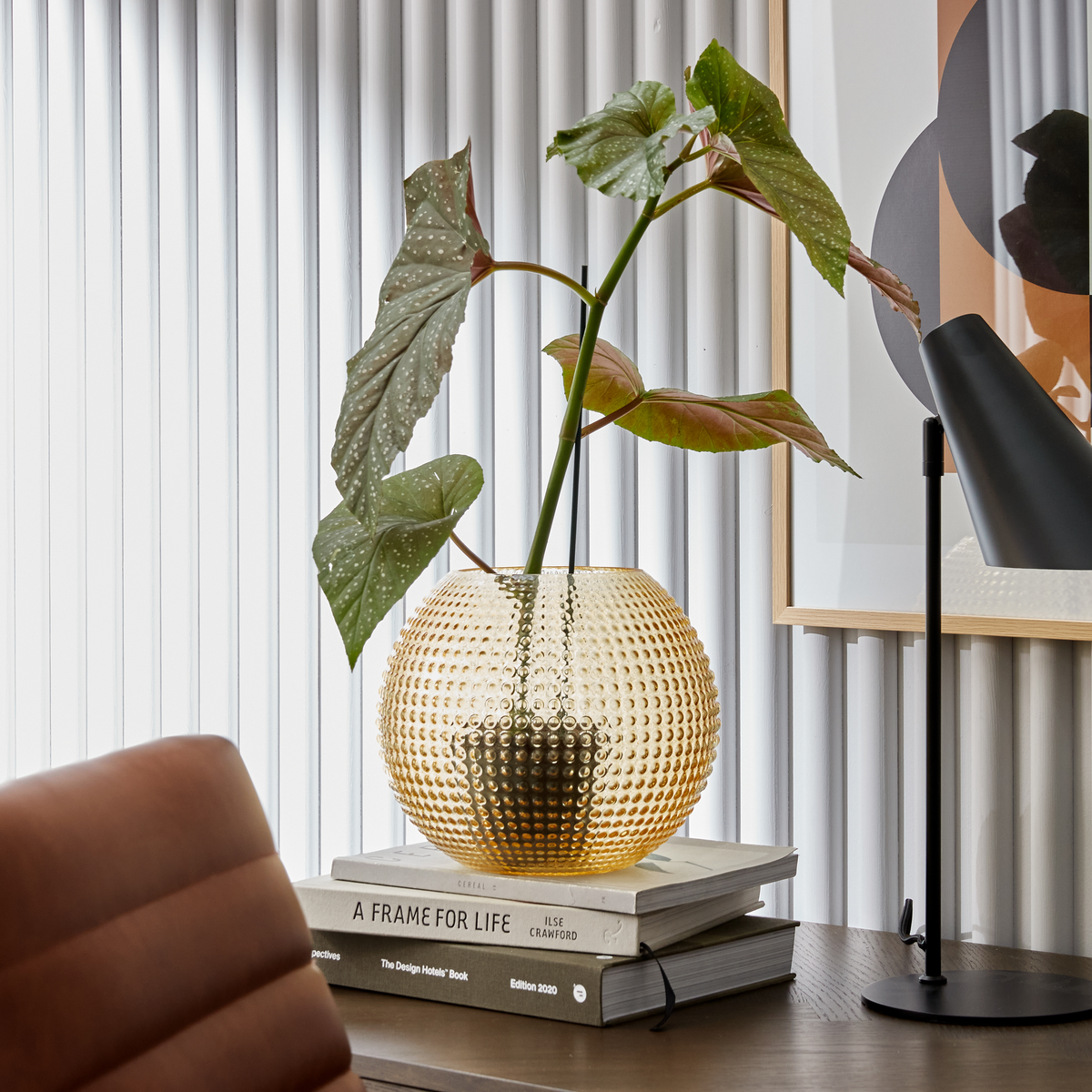 Ravfarvet struktureret glasvase med grøn plante på stablede bøger A Frame for Life af Ilse Crawford og The Design Hotels Book Edition 2020 på et skrivebord ved siden af en sort bordlampe og indrammet kunst.