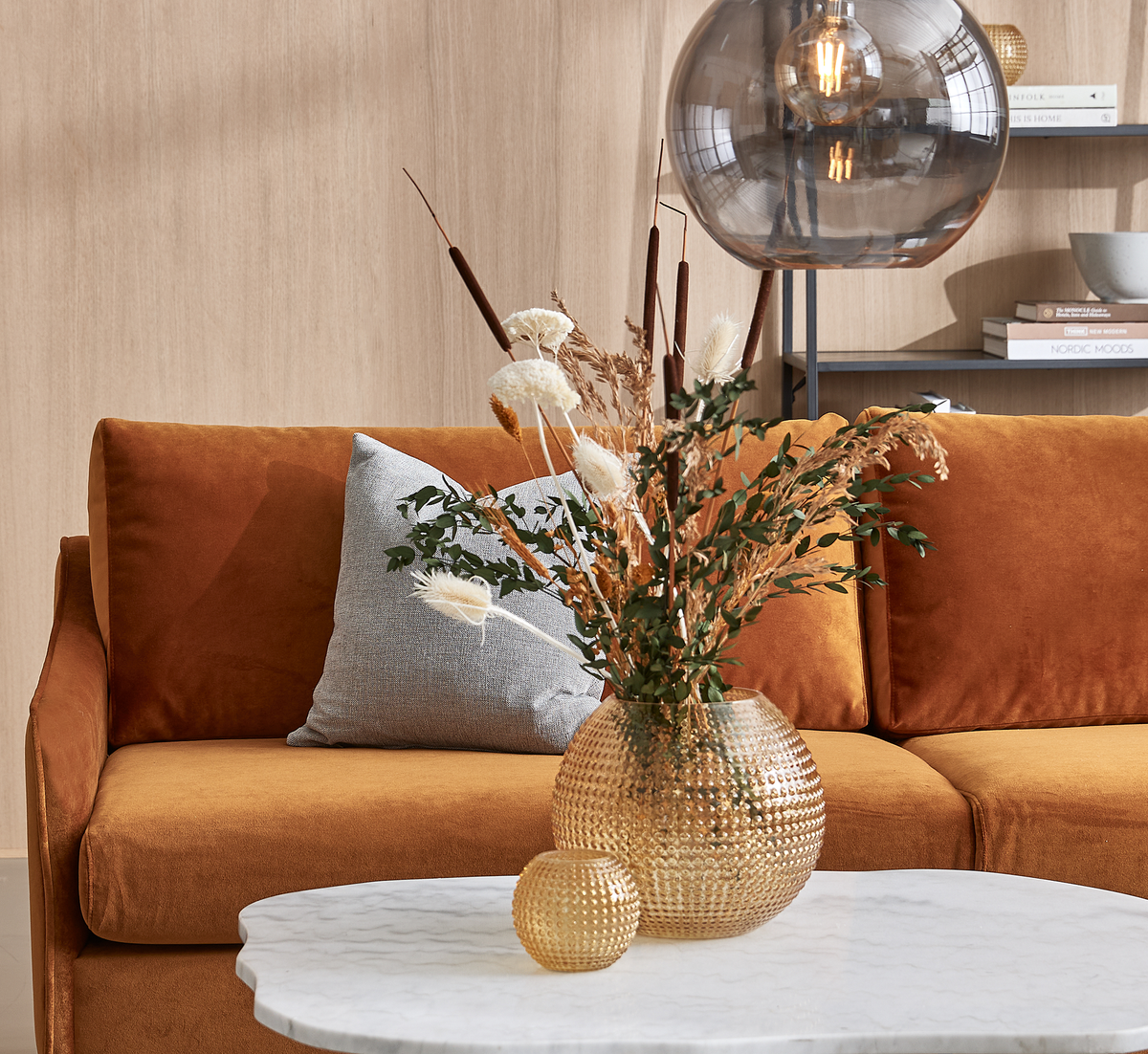 Ravfarvet fløjlssofa med lysegråt pude bag et rundt marmorbord med riflede glasvaser i rav og tørrede blomster; en røgfarvet pendellampe hænger over, og på hylden står bøgerne ”NORDIC MOODS” og ”HOME IS HOME”.
