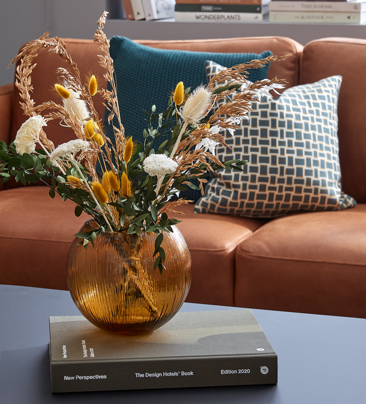 Ravfarvet riflet glasvase med tørrede og grønne grene på sofabord oven på bogen ”New Perspectives – The Design Hotels Book, Edition 2020”, foran en lysebrun sofa med blå mønstrede puder; i baggrunden ses ”WONDERPLANTS” og ”THIS IS HOME”.
