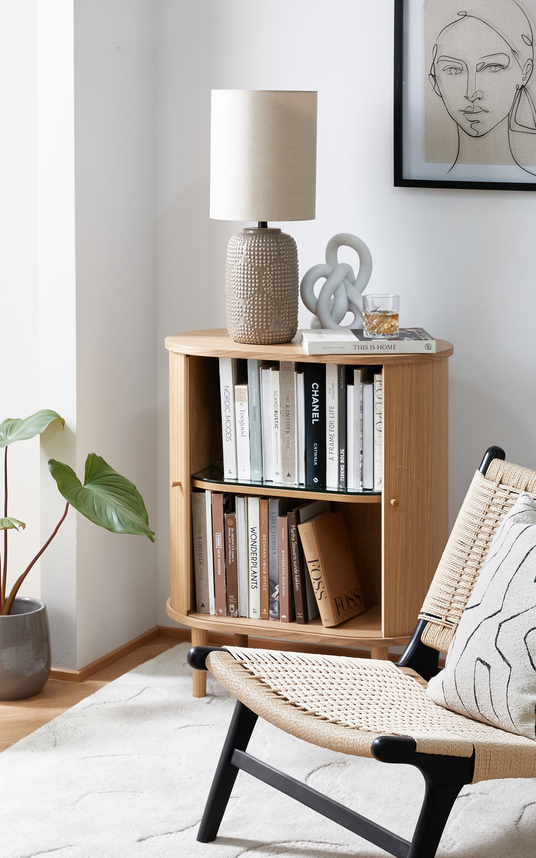 Lys skandinavisk krog med hjørnereol i træ fyldt med designbøger; ovenpå står en beige bordlampe, en skulpturel knude og et glas med drik på bogen ”This Is Home”. En flettet loungestol med pude, en potteplante og et lyst tæppe fuldender scenen; synlige bogtitler omfatter ”CHANEL” og ”Nordic Moods”.
