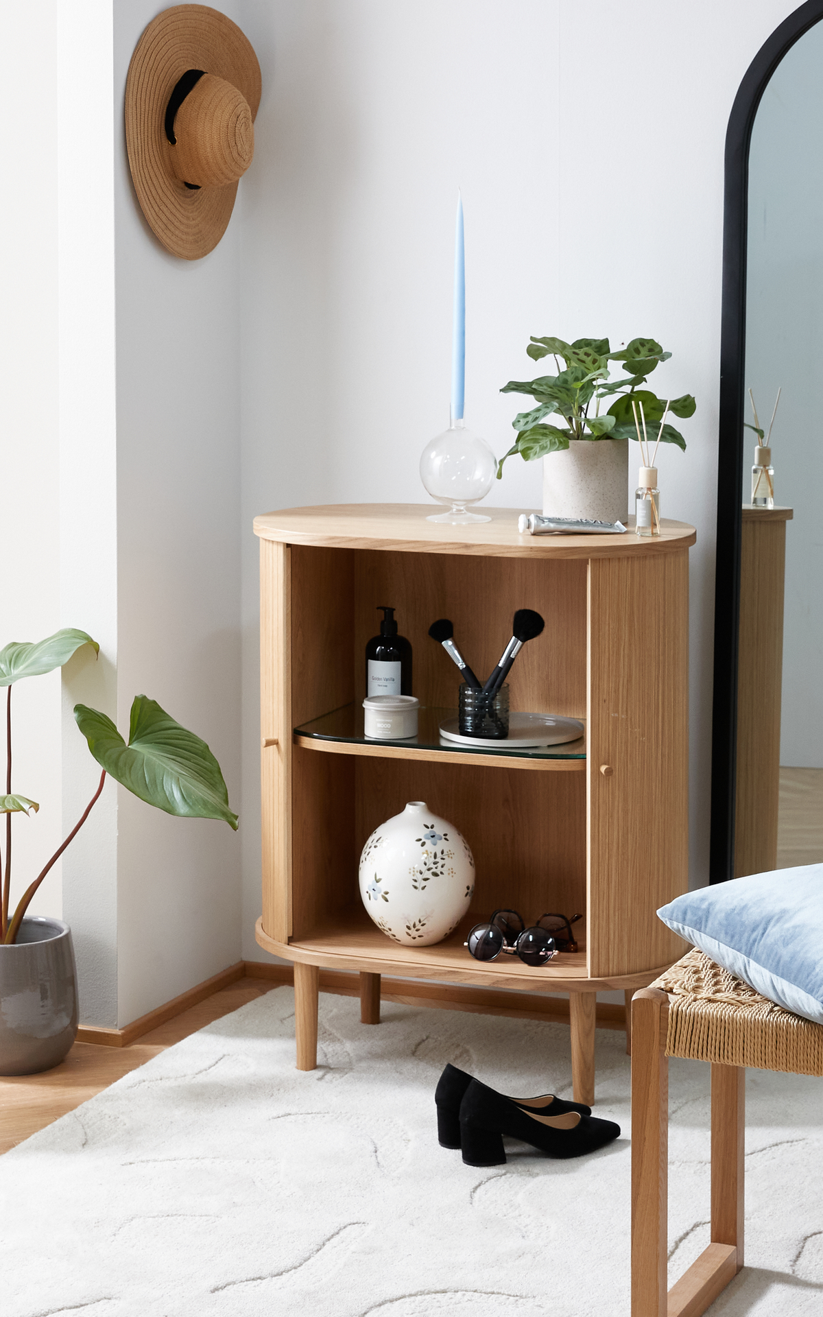 Lyst entréhjørne med lille træskab, spejl med sort ramme, grøn potteplante og lyseblåt stearinlys; sorte pumps på lyst tæppe og stråhat på væggen.