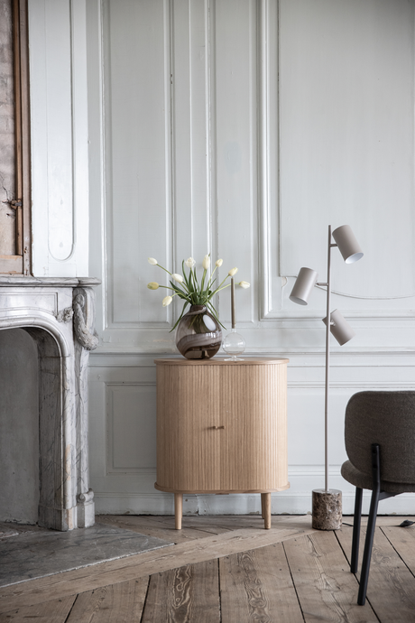 Lys stue med panelvæg, lille skænk i træ med vase med hvide tulipaner og lysestage, ved siden af en moderne gulvlampe og marmorkamin.