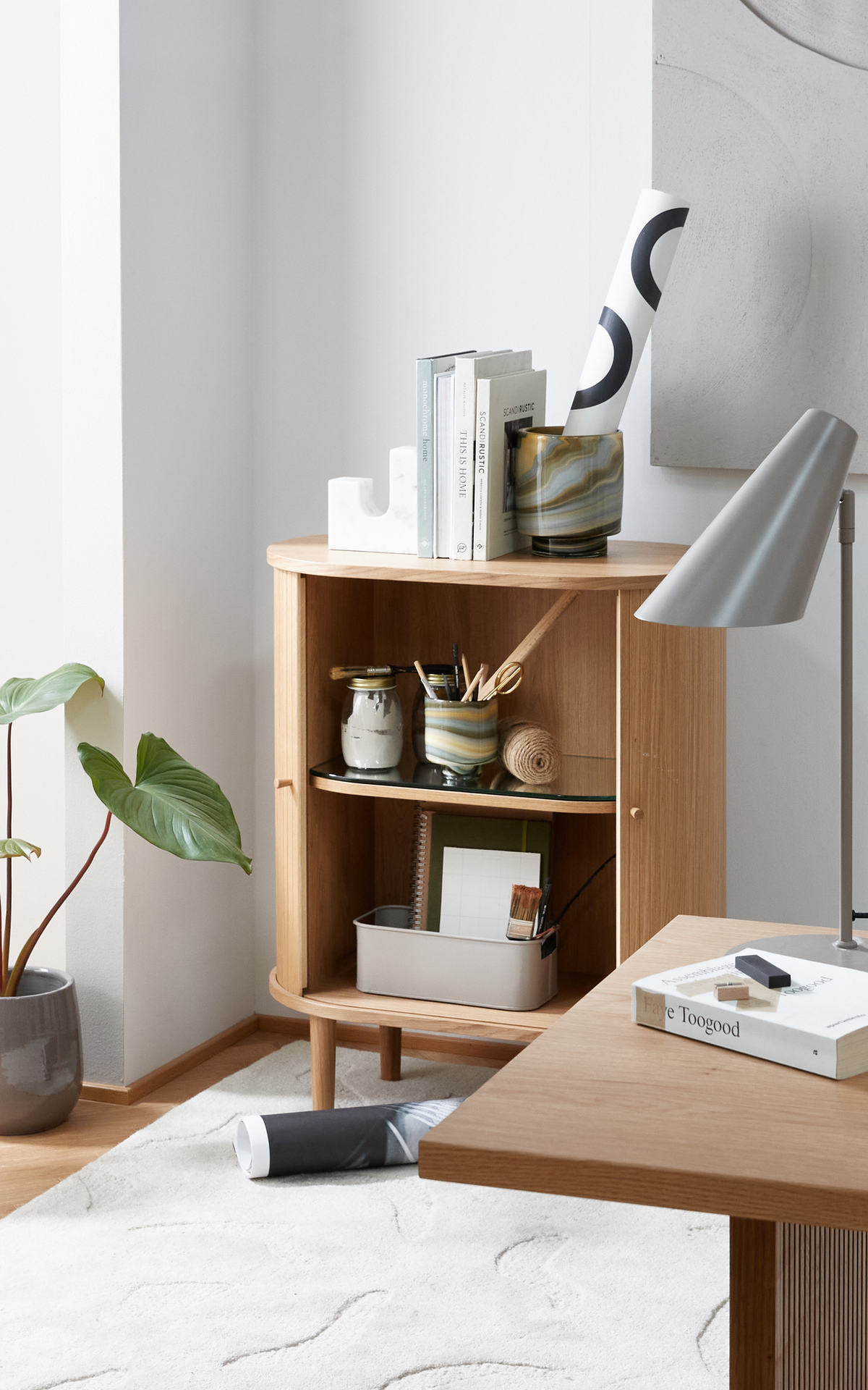 Lyst hjemmekontorhjørne med egetræsbord og rund reol; grå skrivebordslampe, potteplante og lyst tæppe. På hylden står bøgerne ‘SCANDI RUSTIC’ og ‘THIS IS HOME’ samt en rulle med grafisk motiv; på bordet ligger bogen ‘Faye Toogood’.