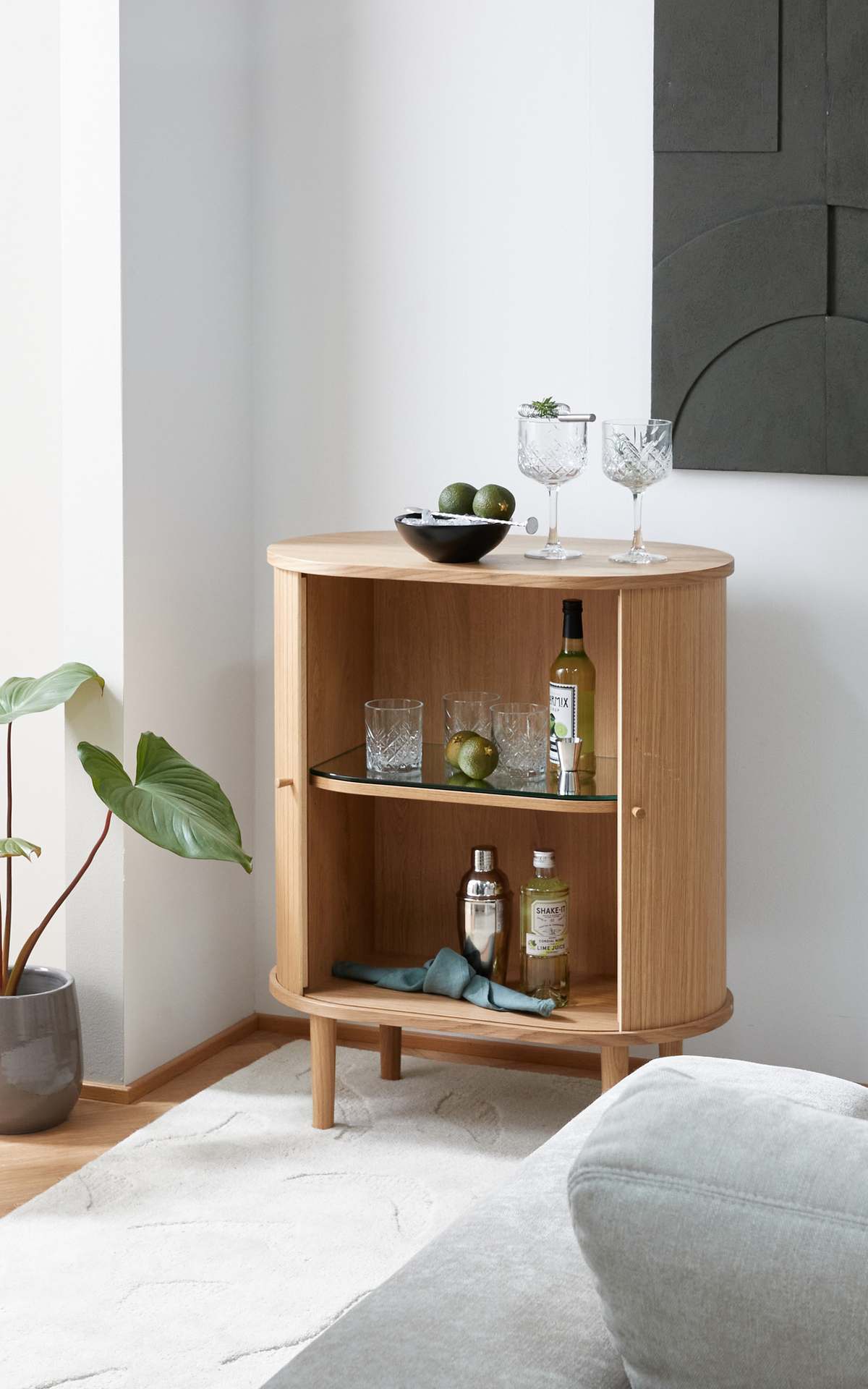 Lille afrundet barskab i egetræ med åbne hylder, glas, shaker, flasker og lime; to krystalglas og en skål med lime ovenpå, ved siden af en potteplante i en lys stue.