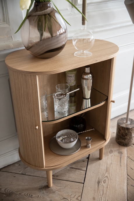 Kompakt egetræsskab som hjemmebar med glas, si, cocktailshaker og en flaske ”SHAKE-IT Lime Juice”; røgfarvet vase med tulipaner ovenpå og en gulvlampe ved siden af.