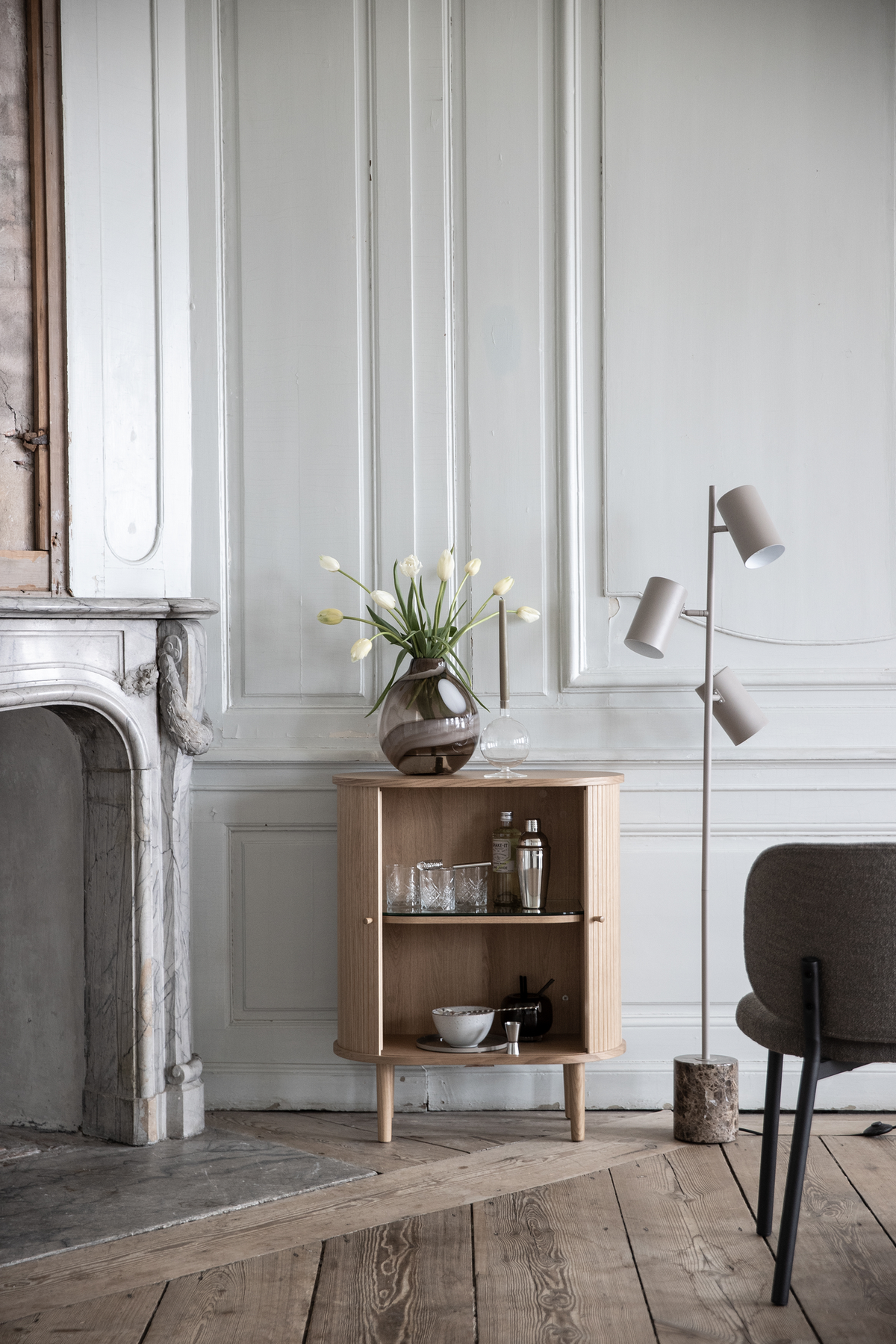 Lys skandinavisk indretning med et lille egetræsskab, en vase med hvide tulipaner og en minimalistisk gulvlampe ved en marmorkamin foran en panelvæg.