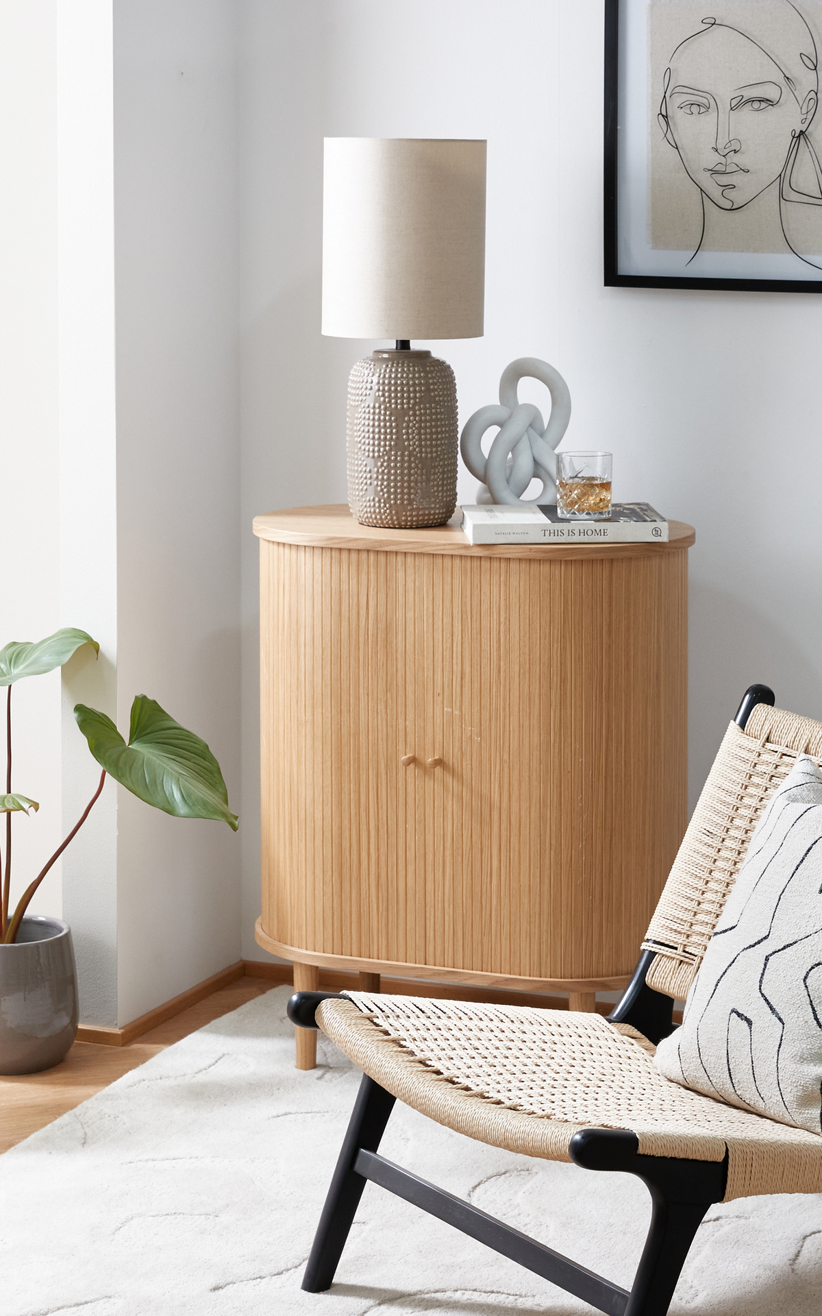 Lyst stuehjørne med halvrundet skab i lys eg, bordlampe med beige skærm, skulpturel knude, et glas på bogen This Is Home, flettet loungestol, potteplante og blød tæppe.