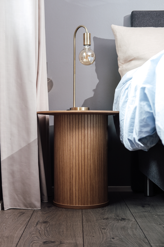 Rundt sengebord i rillet træ med messingfarvet bordlampe med synlig pære ved siden af en redt seng og lyse gardiner.