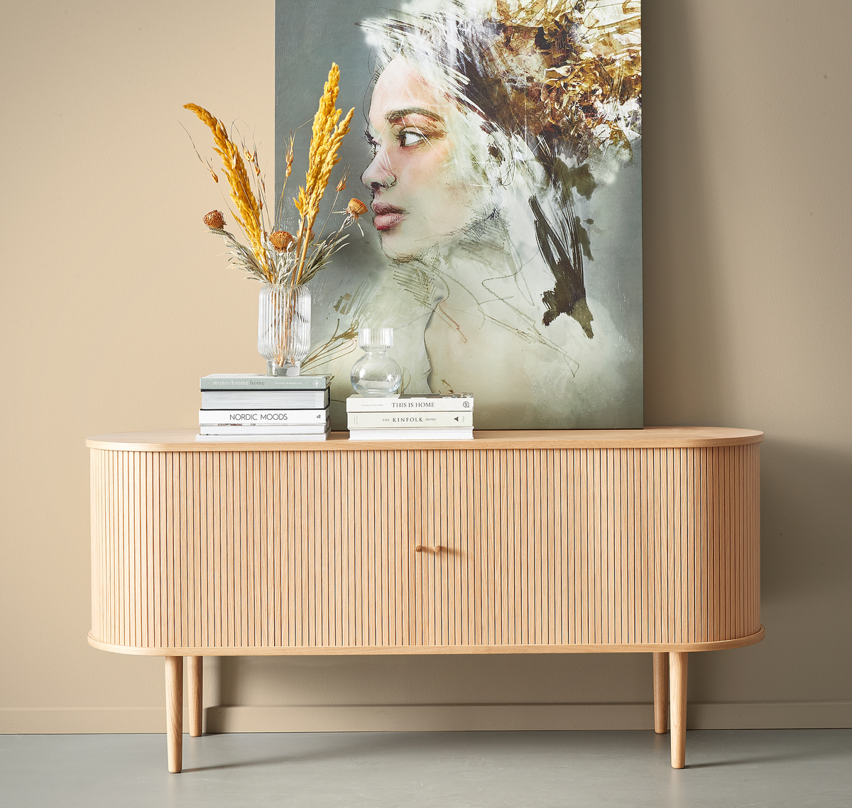 Lyst rillet træskænk med tørrede strå i riflede glasvase, en rund glaskaraffel og bogstakke med titlerne Nordic Moods, This Is Home og Kinfolk foran et abstrakt kunstværk mod en beige væg.