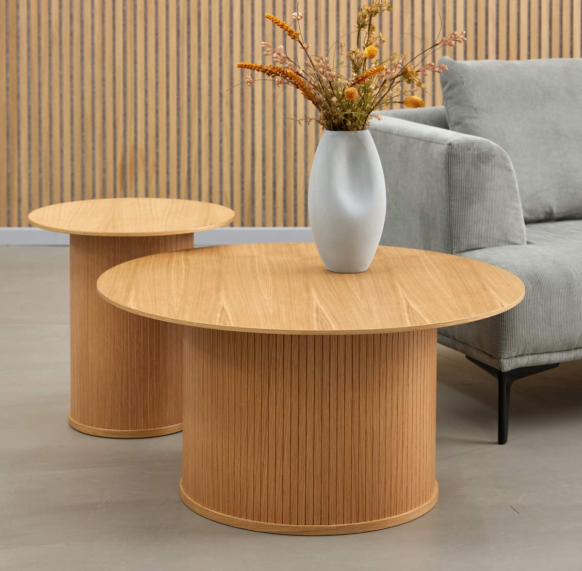 Rundt sofabord og sidebord i lys eg med cylindrisk sokkel; hvid vase med tørrede blomster på bordet og grå sofa i baggrunden i en minimalistisk stue.