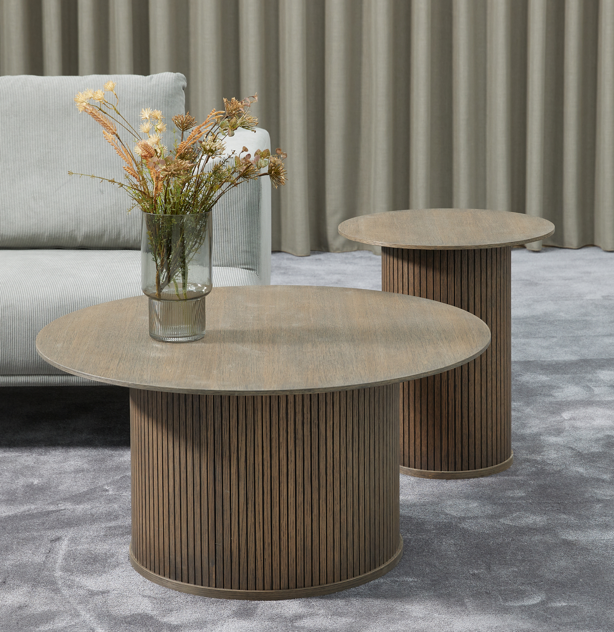 To runde sofaborde i mørkt træ med riflede cylinderfødder; en glasvase med tørrede blomster på det forreste bord, grå sofa, gardiner og gråt tæppe.