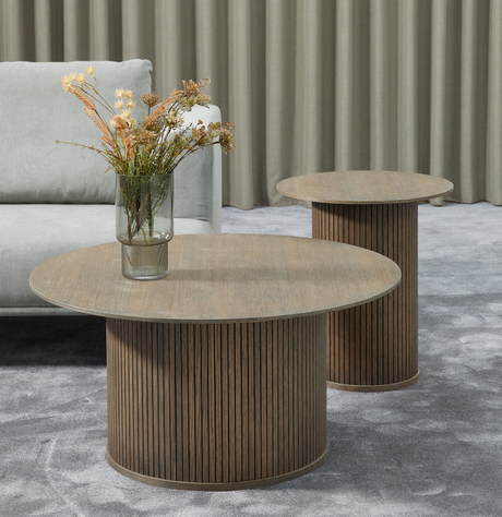 To runde sofaborde i mørkt træ med riflede cylinderfødder; en glasvase med tørrede blomster på det forreste bord, grå sofa, gardiner og gråt tæppe.