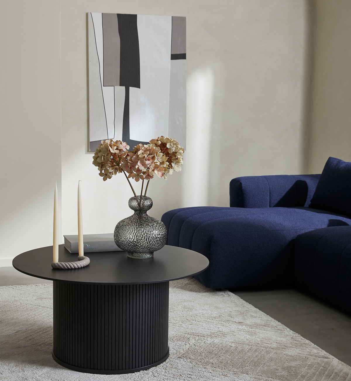 Minimalistisk stue med rundt sort sofabord med riflet sokkel, glasvase med tørrede hortensia, to cremefarvede lys og en Dior-bog; mørkeblå modulsofa og abstrakt vægkunst.