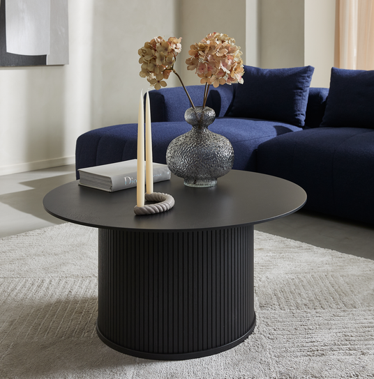 Rundt sort sofabord med rillet søjlefod på lys måtte, stylet med glasvase med tørrede hortensia, to stagelys og en Dior-bog i en moderne stue med mørkeblå sofa.