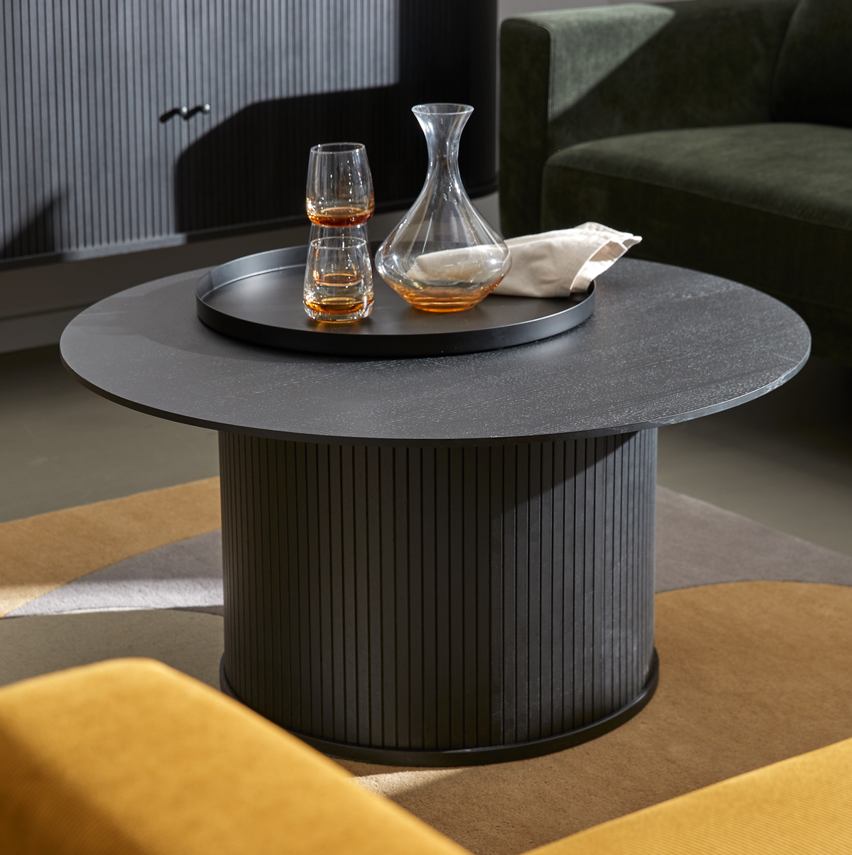Rundt sort sofabord med riflet cylinderfod, med bakke, karaffel og to glas i en stue.