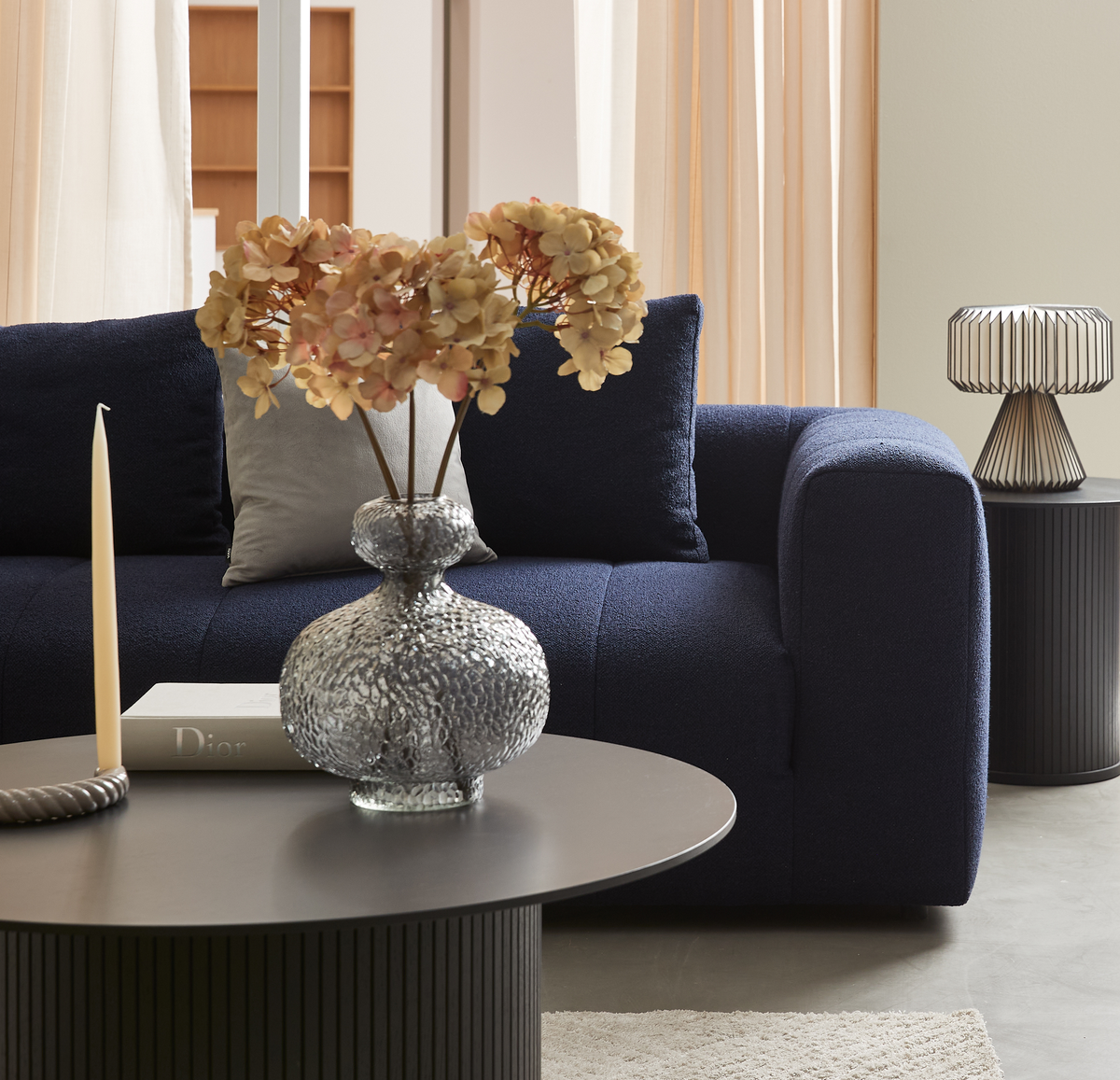 Mørkeblå sofa med puder bag et rundt sofabord med glasvase med tørrede blomster, slank lys og bogen Dior; i baggrunden lette gardiner og en metalbordlampe.