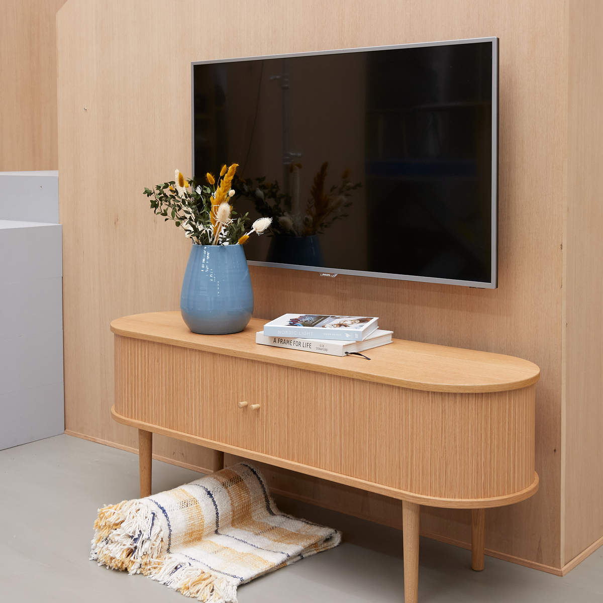 Lys træskænk under vægmonteret tv, med blå vase med tørrede blomster og en bog med titlen ”A FRAME FOR LIFE”; rullet stribet plaid nedenunder.