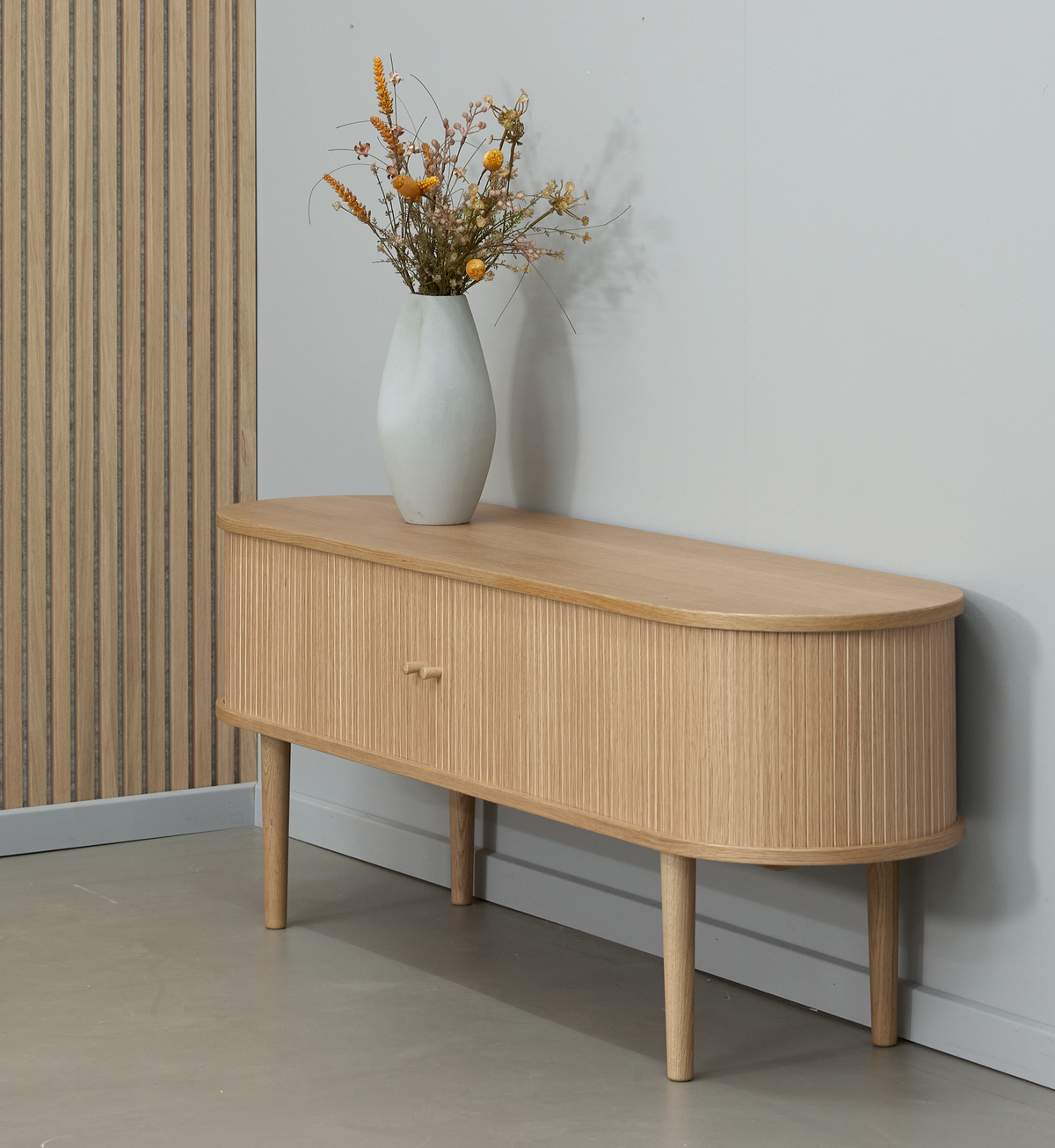 Lav skænk i lyst egetræ med afrundede hjørner og rillet front, vase med tørrede blomster ovenpå mod neutral væg.