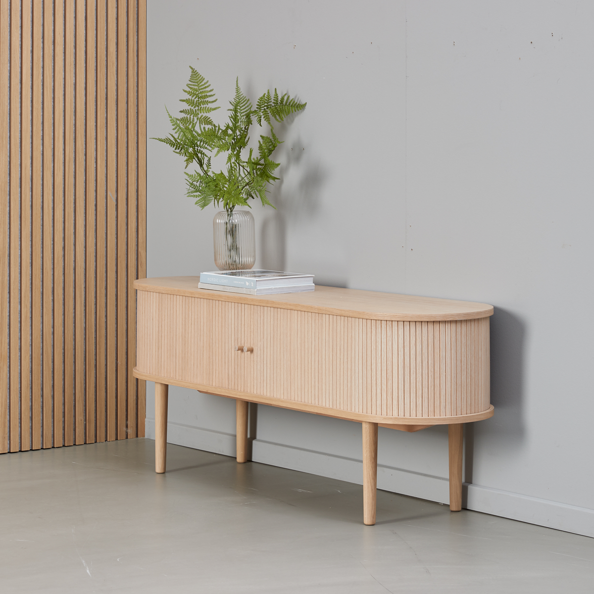 Lyst træsideboard med afrundede hjørner og rillet front, glasvase med bregner og to bøger ovenpå mod lysegrå væg.