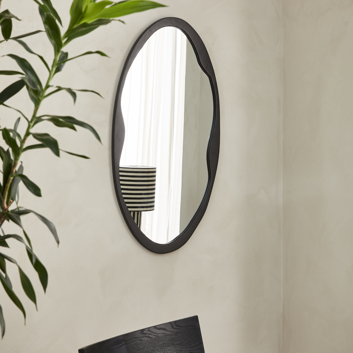 Oval vægspejl med sort, bølgende ramme i et lyst rum; en plante og en stribet lampeskærm reflekteres i glasset.