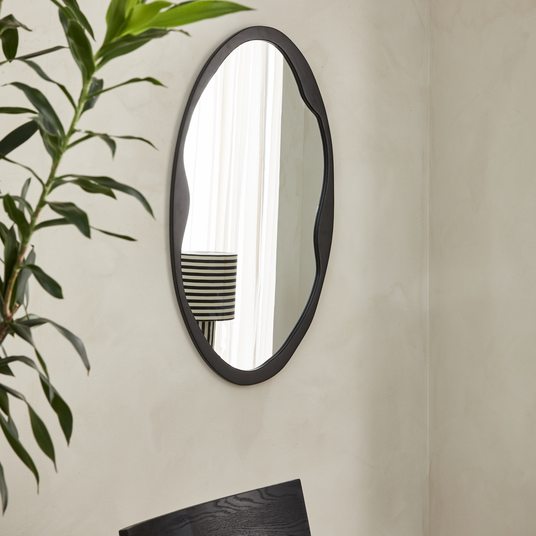 Oval vægspejl med sort, bølgende ramme i et lyst rum; en plante og en stribet lampeskærm reflekteres i glasset.