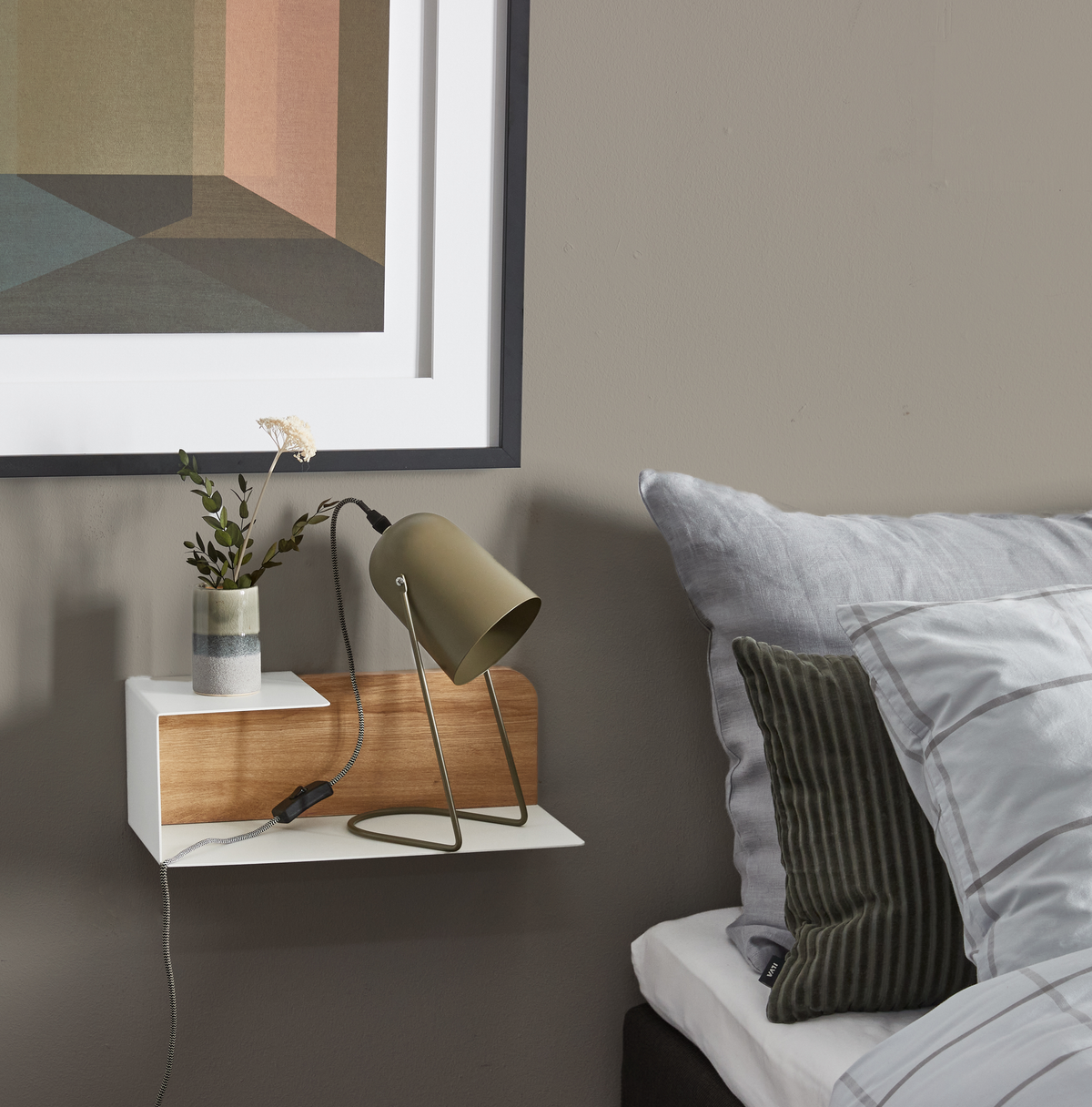 Soveværelsesdetalje med væghylde i hvid og træ, olivengrøn bordlampe og lille vase med gren; gråt sengetøj og indramet geometrisk plakat ovenover.