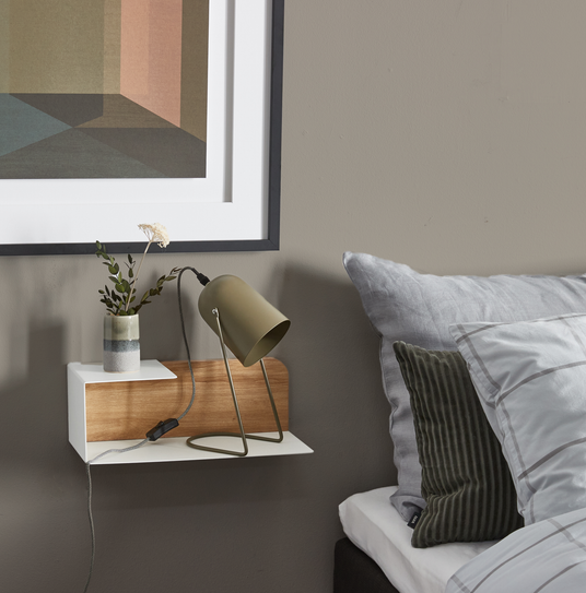 Soveværelsesdetalje med væghylde i hvid og træ, olivengrøn bordlampe og lille vase med gren; gråt sengetøj og indramet geometrisk plakat ovenover.