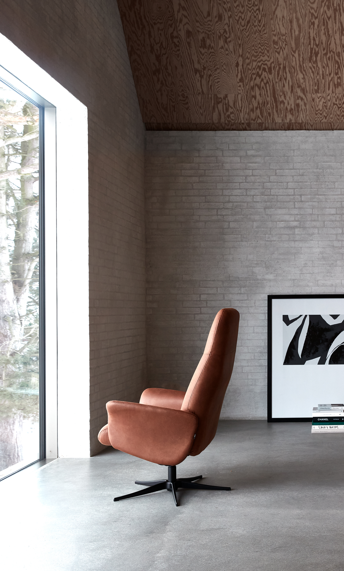 Minimalistisk stue med en brun drejestol ved et stort vindue, lys murstensvæg, indrammet abstrakt kunst og en bogstak med "CHANEL".