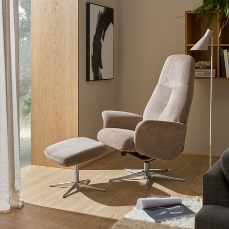 Beige hvilestol med matchende skammel på metalsokkel i en lys stue ved vinduet, med gulvlampe, vægkunst og åben reol i dagslys.