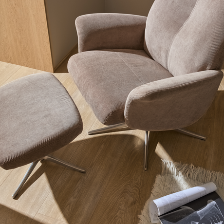 Beige lænestol i fløjl med matchende skammel på trægulv, begge med metalben; solbelyst hjørne med lyst rygtæppe og et opslået magasin på gulvet.