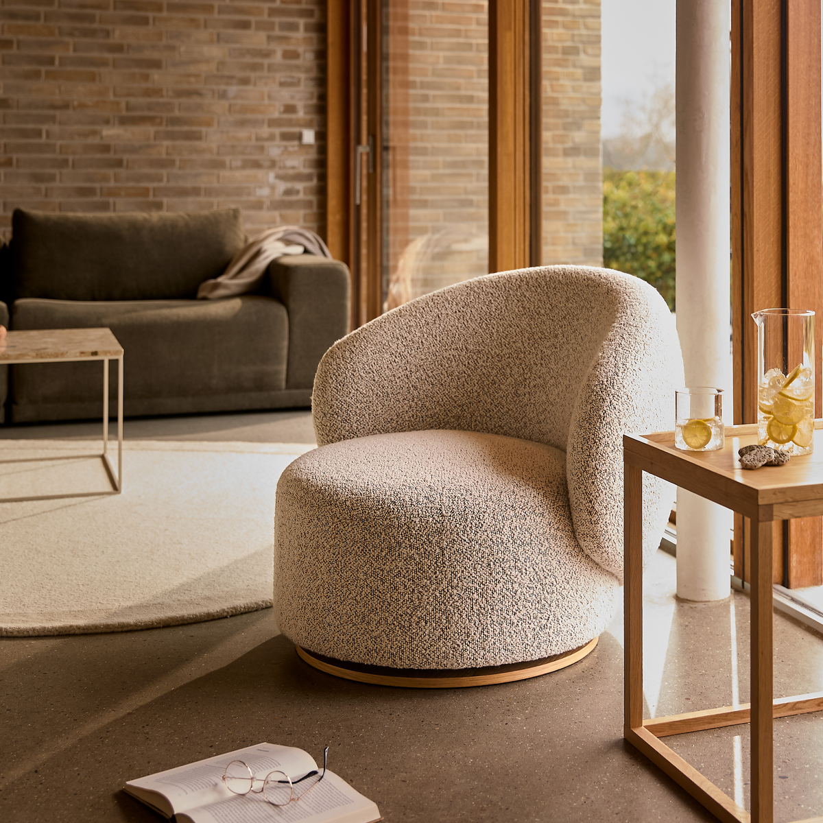 Bouclé drejestol i en varm, minimalistisk stue med sofa, lyst tæppe og træbord med glas og karaffel med is; sollys fra store vinduer.