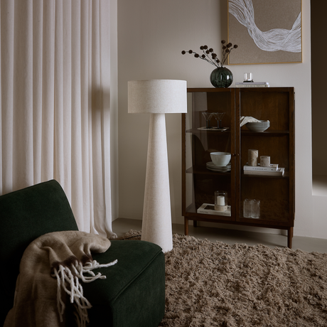 Hyggeligt hjørne af stuen med mørkegrøn loungestol med beige plaid, høj hvid gulvlampe, shaggy tæppe og vitrineskab i mørkt træ med service, bøger, vase og lys.