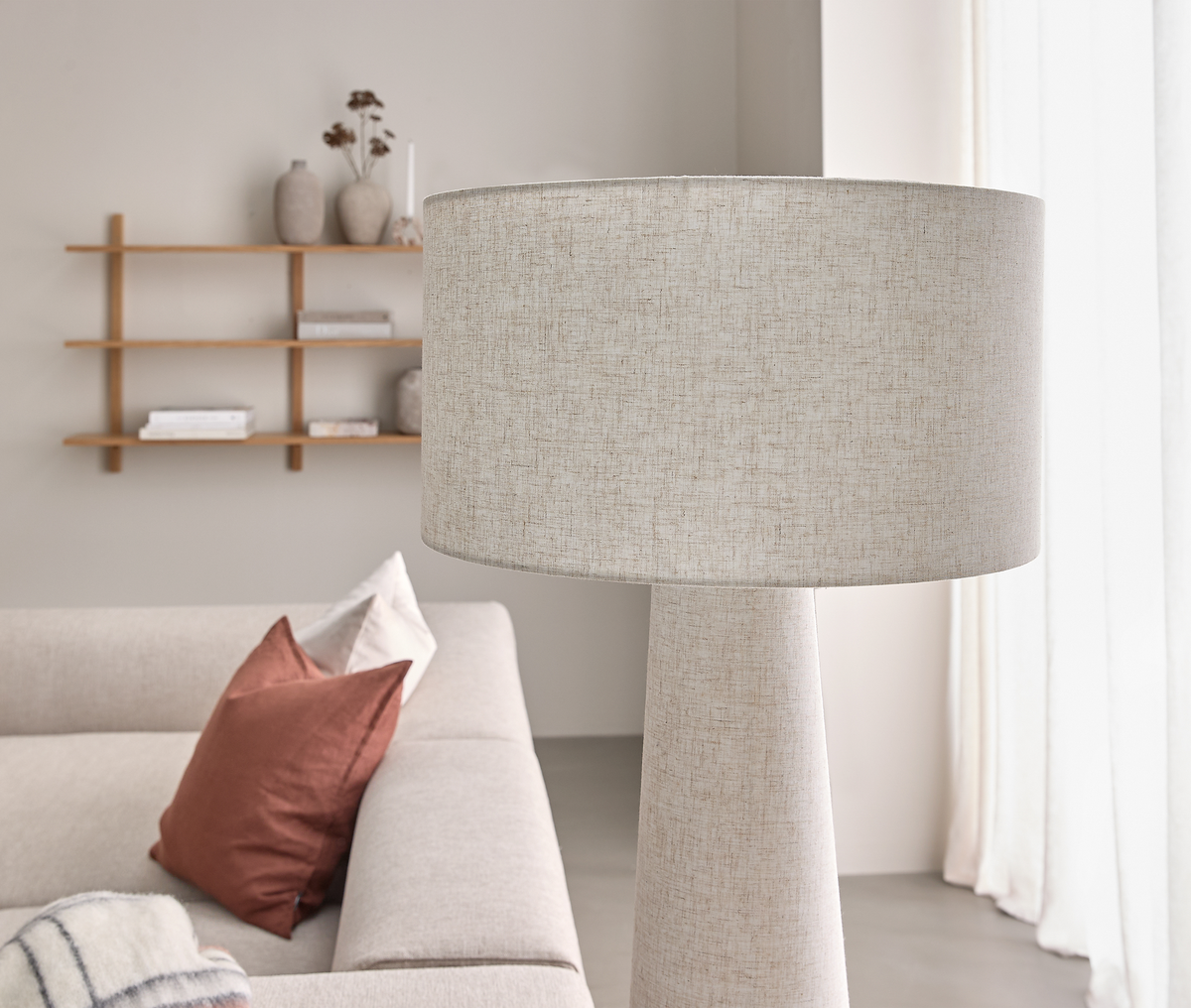 Beige gulvlampe i stof ved siden af en lys sofa med terrakottapuder; hylde og lyse gardiner i en skandinavisk stue.