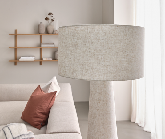 Beige gulvlampe i stof ved siden af en lys sofa med terrakottapuder; hylde og lyse gardiner i en skandinavisk stue.