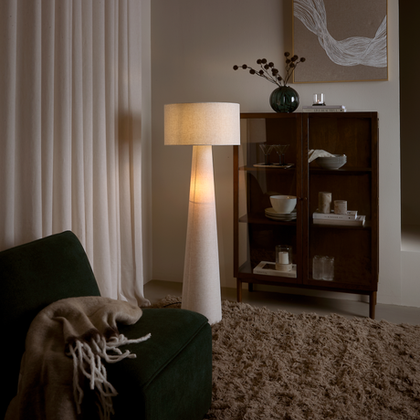 Mørkegrøn lænestol med beige plaid, tændt gulvlampe ved et vitrineskab; tykt shaggy tæppe og lyse gardiner i en varm, hyggelig stue.