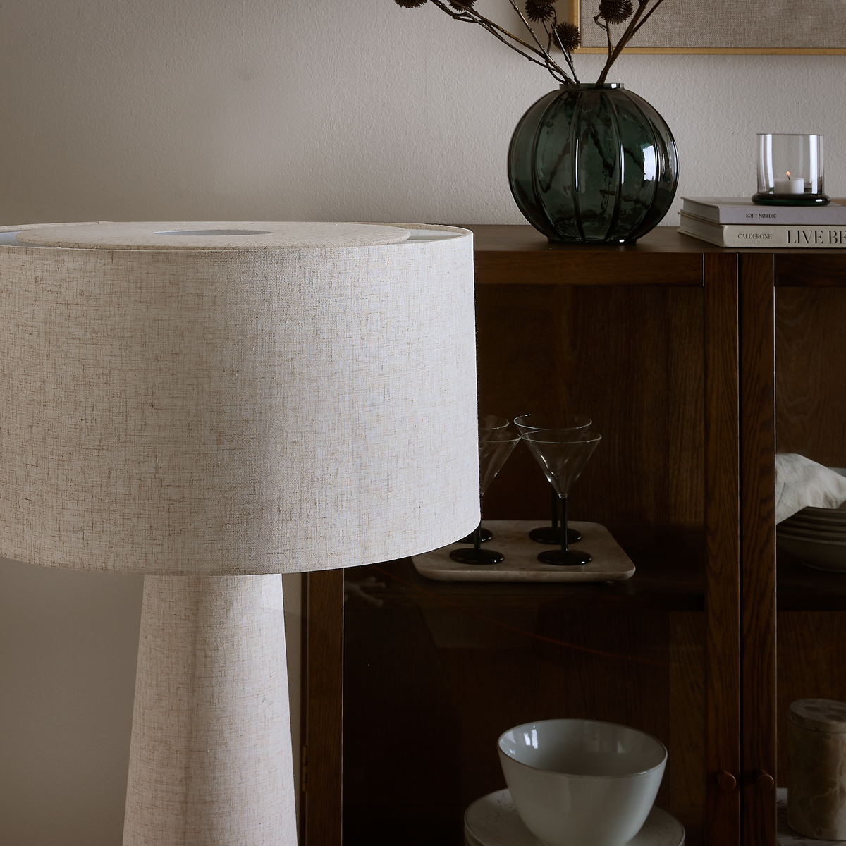 Beige tekstil-gulvlampe ved siden af et mørkt vitrineskab; grøn riflet glasvase med tørrede grene og en tænd fyrfadsstage på bøgerne ”Still Nordic” og ”Live Beautiful”; martiniglas og skåle bag glaslågen.